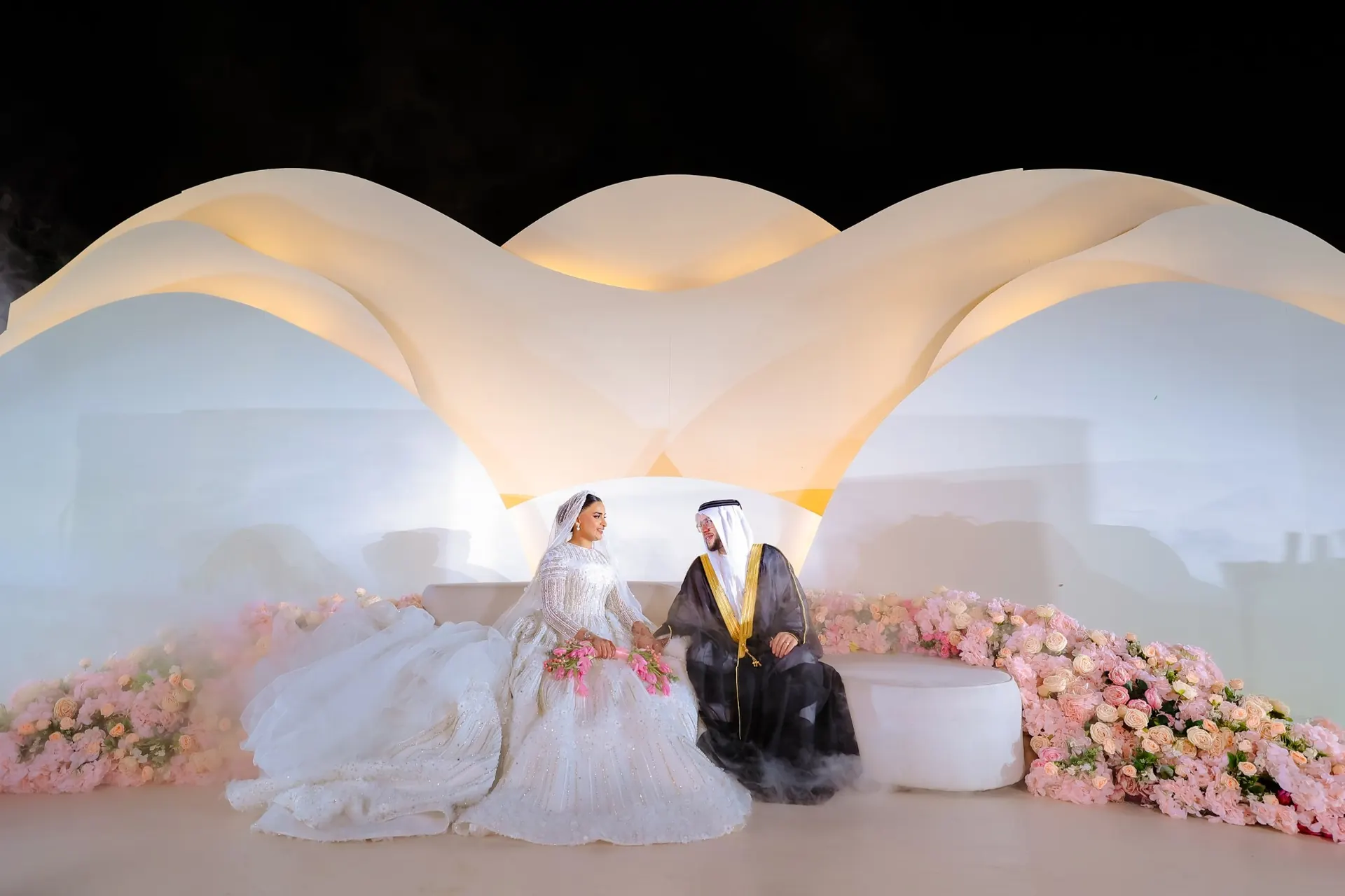 Bride and groom seated on a sculpted white stage with pink floral arrangements at a Dubai desert wedding